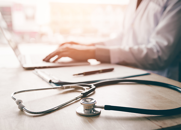 doctor in white coat typing on laptop in background stethoscope on desk in foreground